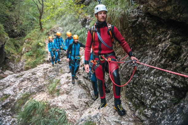 In a canyoning experience, a diverse group of friends, guided by a skilled Caucasian instructor, climbs through challenging terrain in neoprene suits and safety helmets.