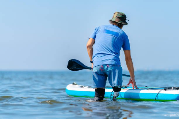 An amputee man is getting on a stand up puddling surfing board in the sea.